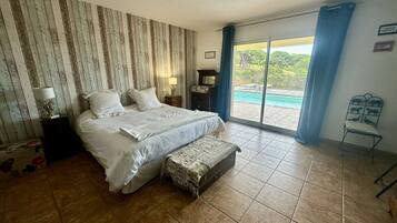 Une chambre avec un grand lit, un banc, une table de chevet, une lampe et une vue sur une piscine, visible à travers une porte coulissante.