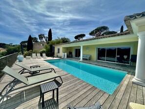 A swimming pool with a wooden deck, lounge chairs, and a building with a tiled roof.
