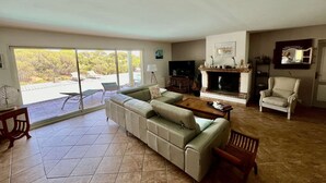 A spacious living room with a sectional sofa, a television, a fireplace, and a balcony with a view of trees. - VILLA WITH SWIMMING POOL TO RENT FOR 6 PEOPLE, QUIET AREA 1.2 KM FROM THE BEACH (STE MAXIME)