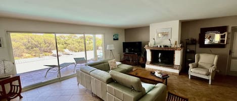 Un salon spacieux avec un canapé d’angle, un téléviseur, une cheminée et un balcon offrant une vue sur les arbres.