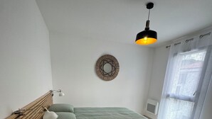 A bedroom with a bed, a bedside table, a lamp, a wall clock, and a window with curtains. - Cosy apartment in new residence  (Propriano)