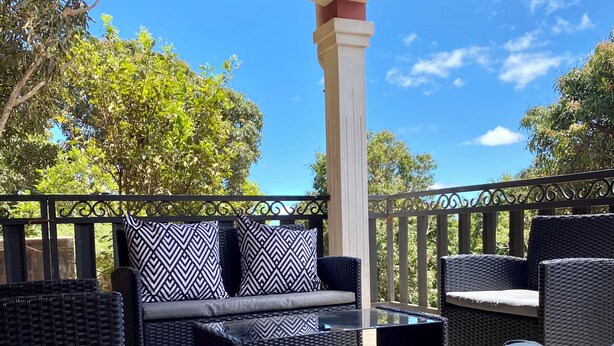 An outdoor patio with wicker furniture, including a sofa and chairs, under a covered area with a view of trees and a clear sky.