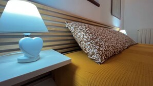 Un lit bien fait, avec un oreiller à motifs et une lampe de chevet en forme de cœur posée sur une table de chevet blanche.