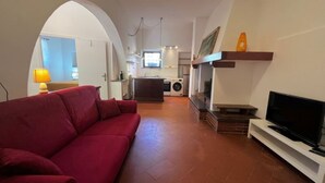 Un salon avec un canapé rouge, un téléviseur posé sur un meuble, une cheminée et, au fond, une cuisine.