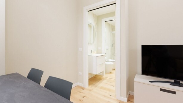 A modern room with a dining table, chairs, a TV, and a bathroom visible through an open door. - GuestHost - Primaticcio M1 Modern and Bright Apartment (Milano)