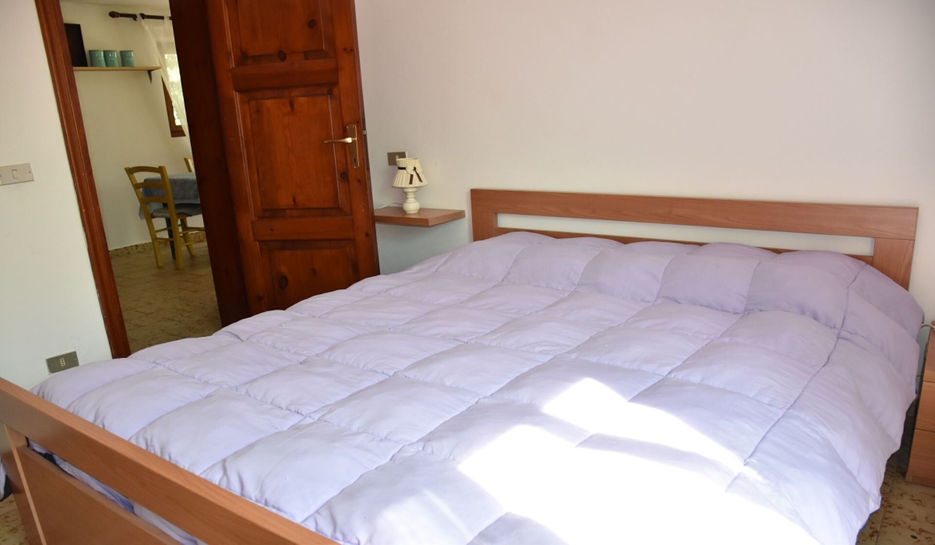 Een enkel bed met een houten hoofdbord en een lichtgekleurd dekbed in een kamer met een houten deur en een klein nachtkastje met een lamp.