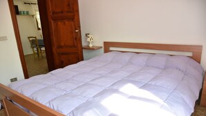Un lit simple avec une tête de lit en bois et une couette de couleur claire, dans une pièce dotée d’une porte en bois et d’une petite table de chevet avec une lampe.