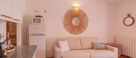 Une cuisine moderne avec un réfrigérateur blanc, un four à micro-ondes et un plan de travail en granit. On y trouve un canapé beige avec des coussins et une petite table d’appoint. Un élément décoratif mural et un anneau lumineux sont également visibles.
