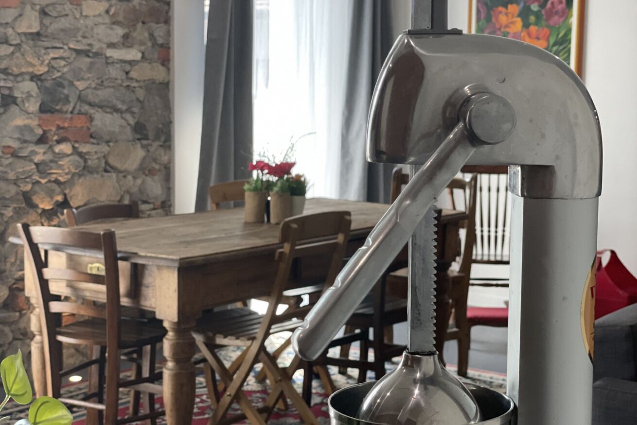 A rustic dining area with a wooden table and chairs, a stone wall, and a metal object in the foreground.