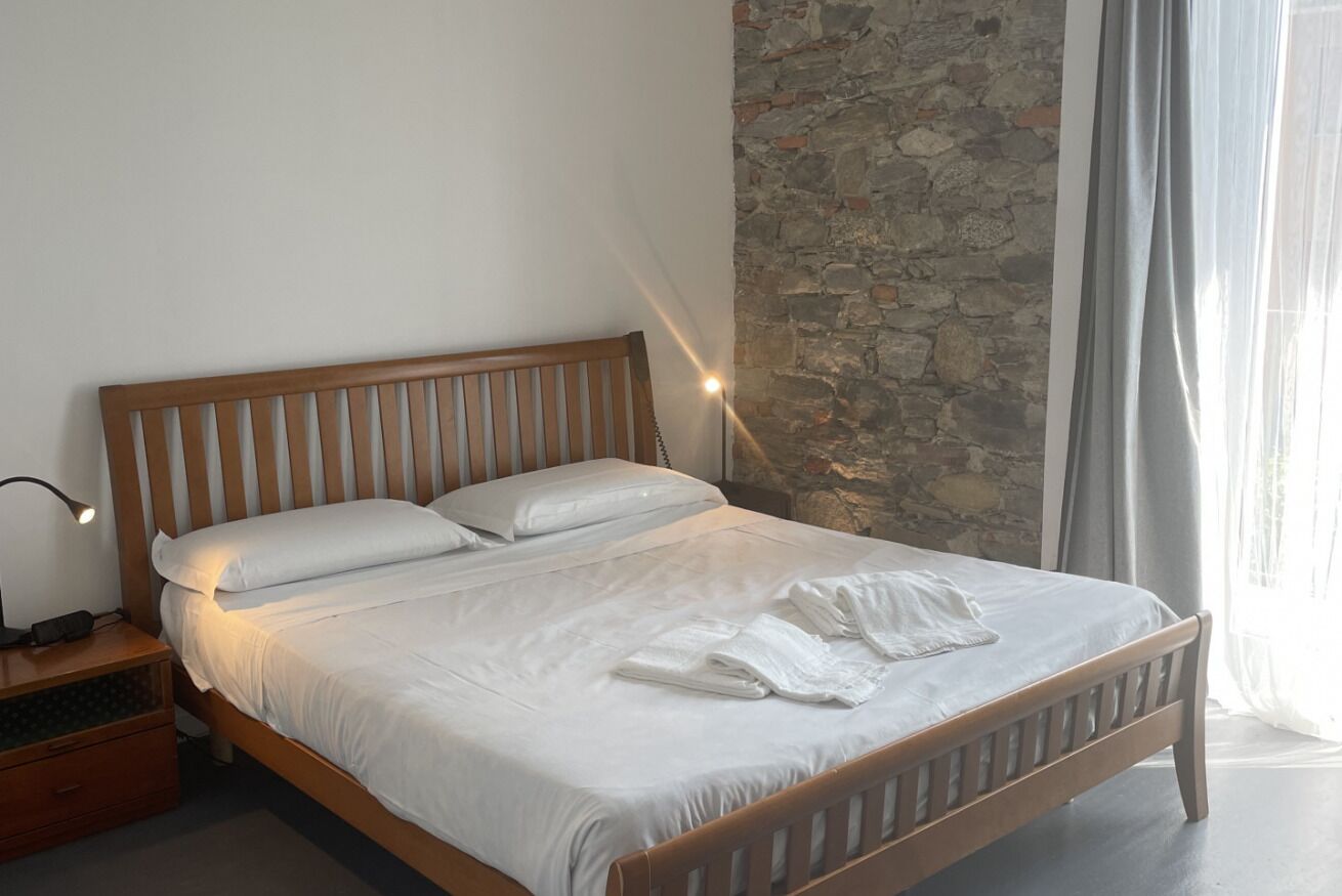 A neatly made bed with white linens and towels, a wooden headboard, a nightstand with a lamp, and a stone wall behind the bed.