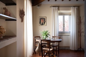 Zona de menjador amb taula, cadires, un vas amb flors, una fotografia amb marc i una finestra amb cortines.