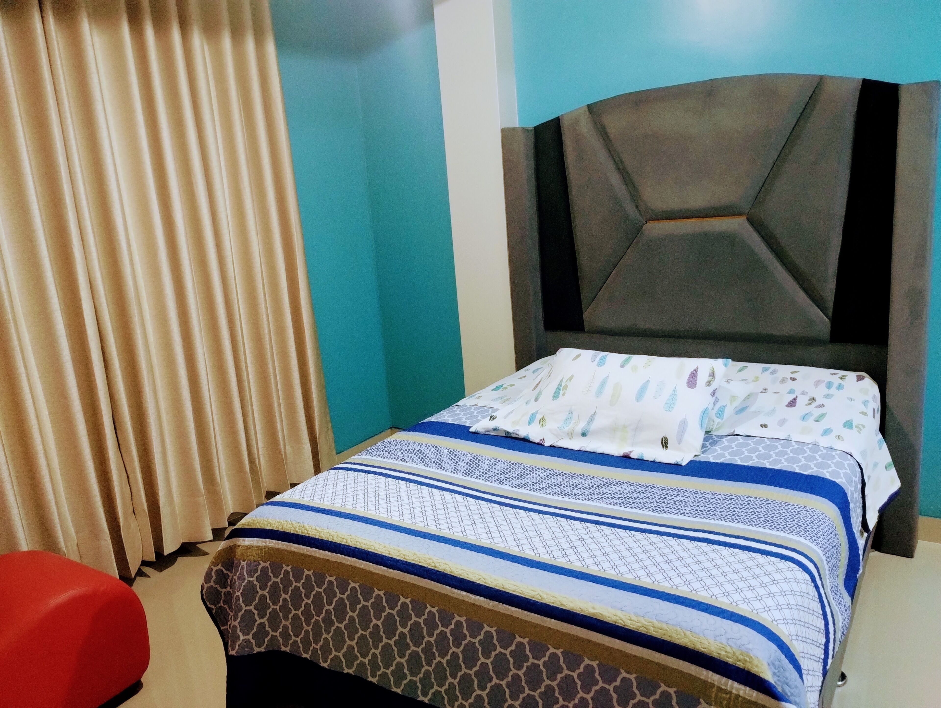 Una cama bien hecha con ropa de cama estampada, una silla roja y una ventana con cortinas.