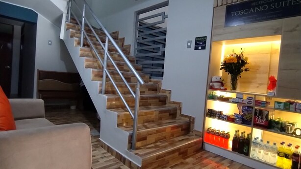 Un escalier avec des marches en bois et des rampes en métal, un comptoir d’accueil avec une enseigne indiquant « RÉCEPTION TOSCANO SUITES », et un présentoir de boissons en bouteille.