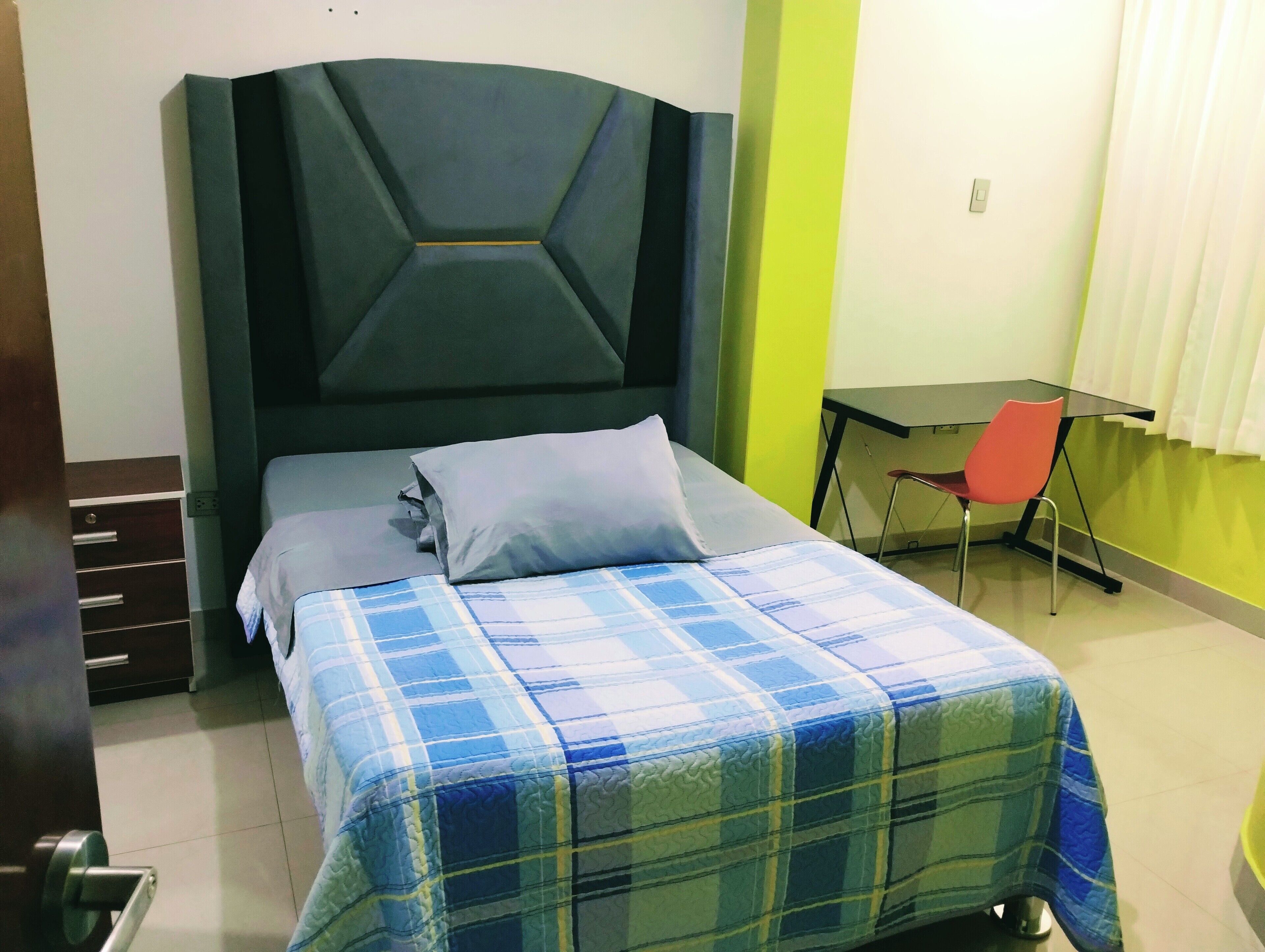 A bed with a plaid bedspread, a bedside table, a chair, and a desk with a chair.