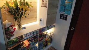 Un comptoir d’accueil avec une enseigne indiquant « Toscano Suites » et un code QR pour l’enregistrement. On y trouve un vase avec des fleurs et un ours en peluche. En dessous, des étagères sont remplies de bouteilles d’eau et de collations.