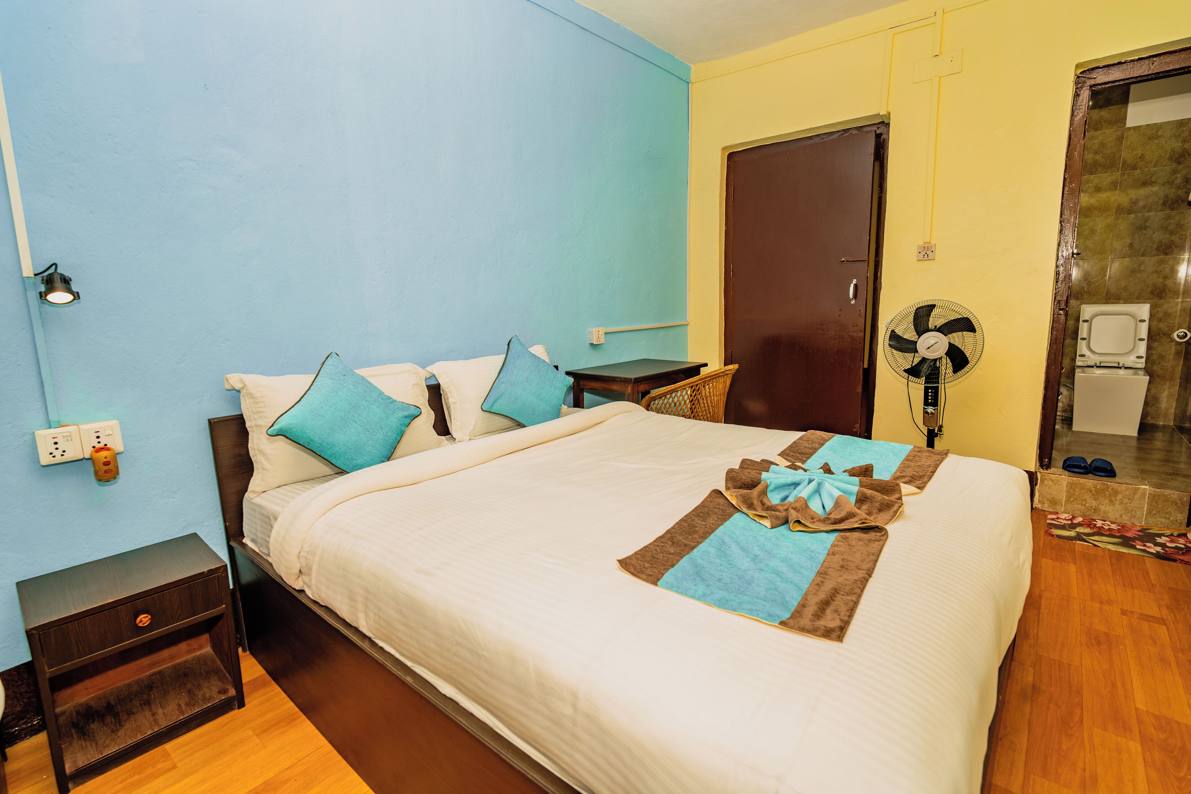 A neatly made bed with a towel and a fan, a nightstand, a wooden floor, and a door leading to a bathroom.