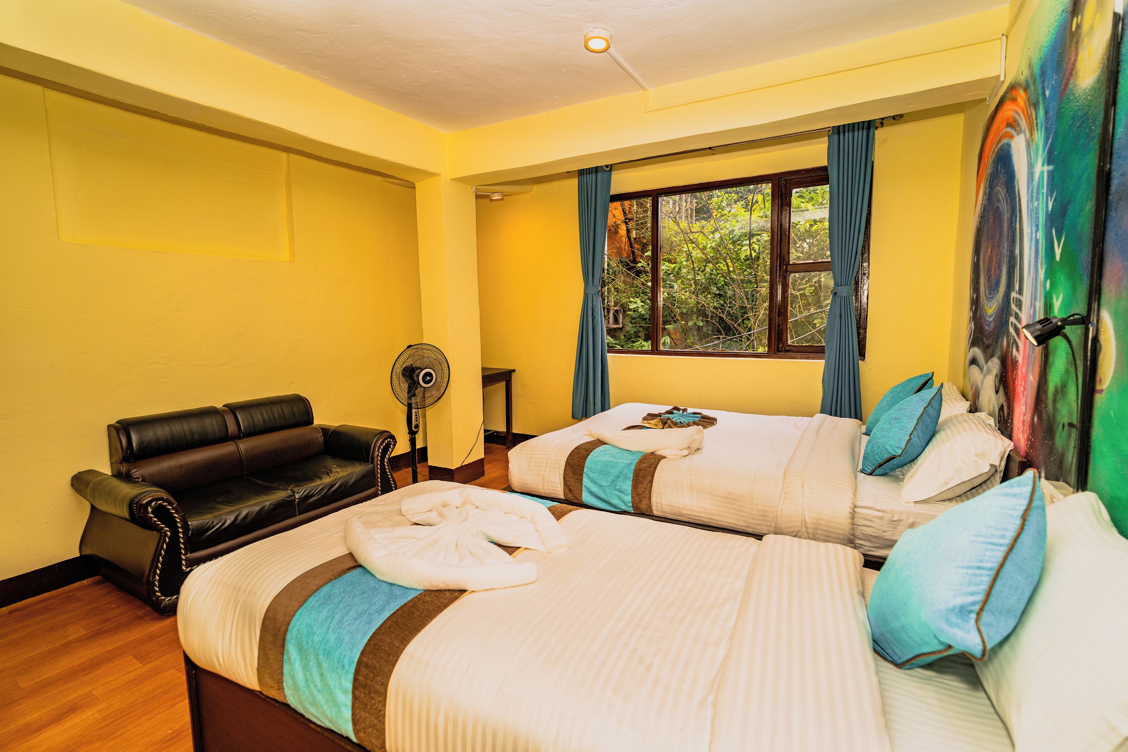 A hotel room with two beds, a sofa, a standing fan, and a window with curtains.