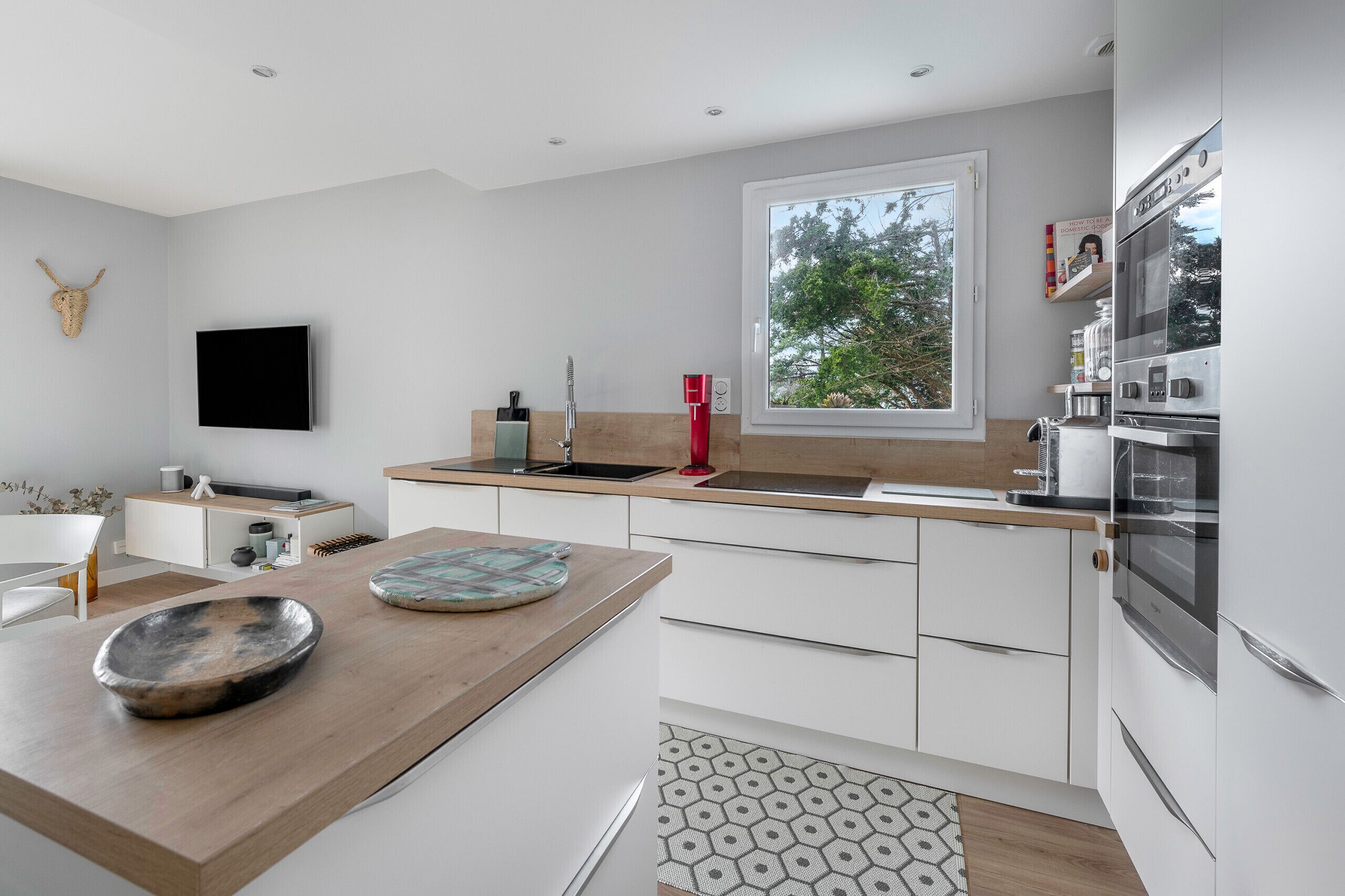 A modern kitchen with white cabinets, a wooden countertop, a large window, and a mounted television.