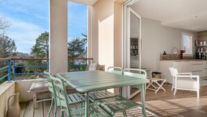 Ein Balkon mit Tisch und Stühlen, eine Küchenzeile mit weißen Schränken und einem Spülbecken sowie Blick auf Bäume und Gebäude.
