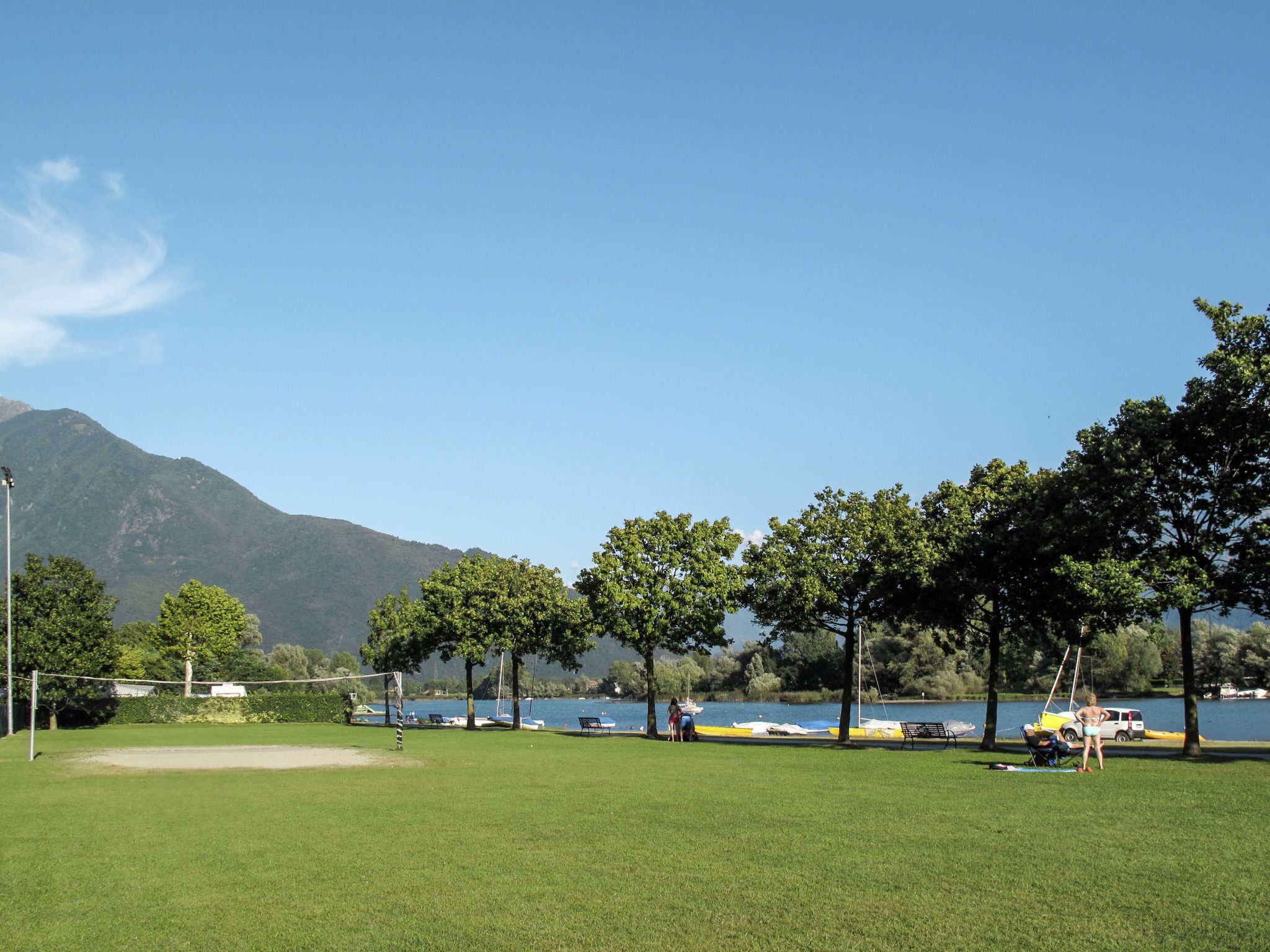 A park with a volleyball net, trees, and a lake with boats.