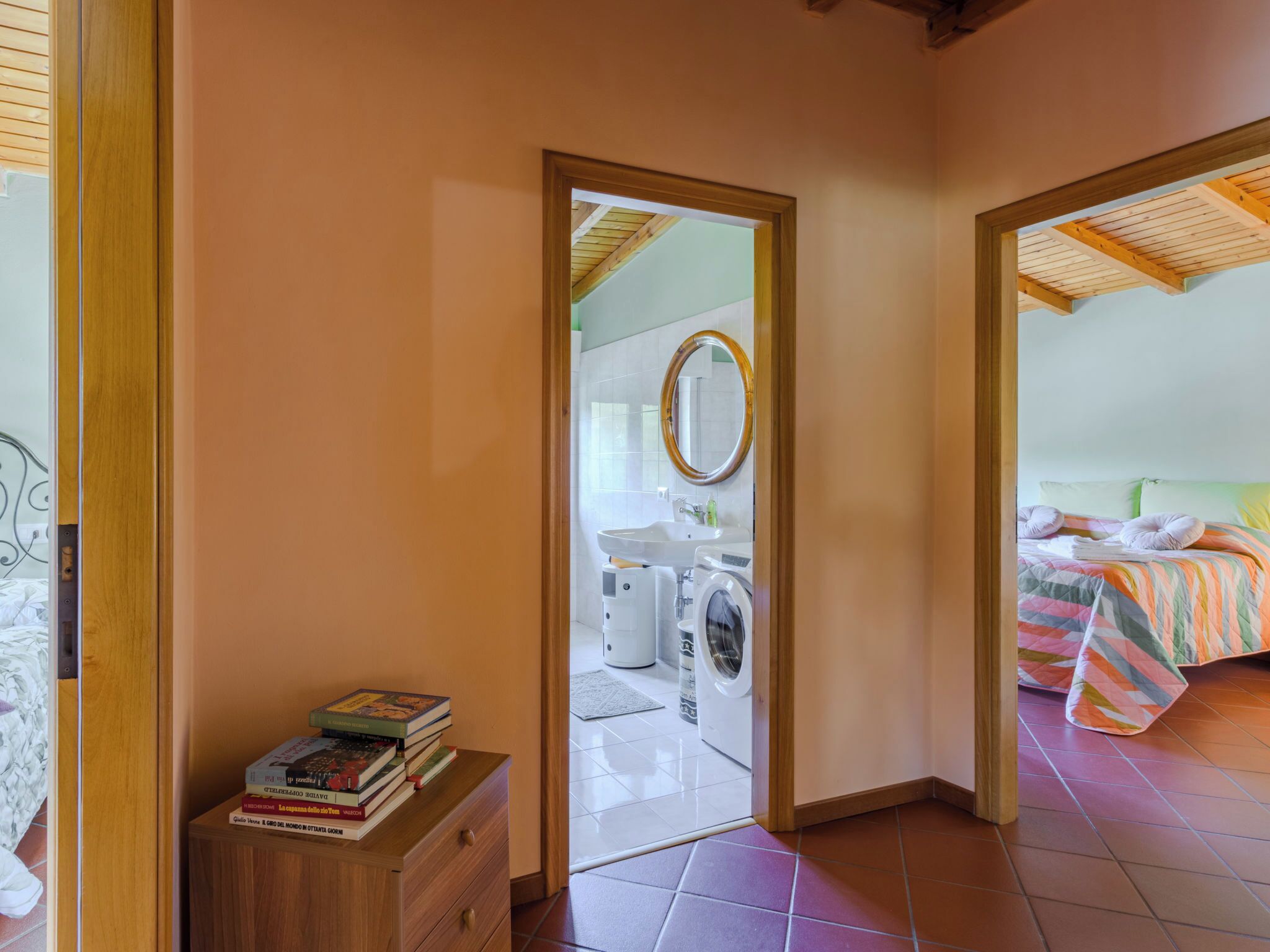 Een kamer met een houten garderobe, een klein nachtkastje met boeken, een bed met een kleurrijke sprei, en een wasmachine in de aangrenzende kamer.