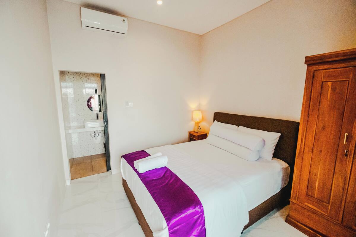 A hotel room with a bed, a wooden wardrobe, a nightstand with a lamp, and a bathroom visible through an open door.