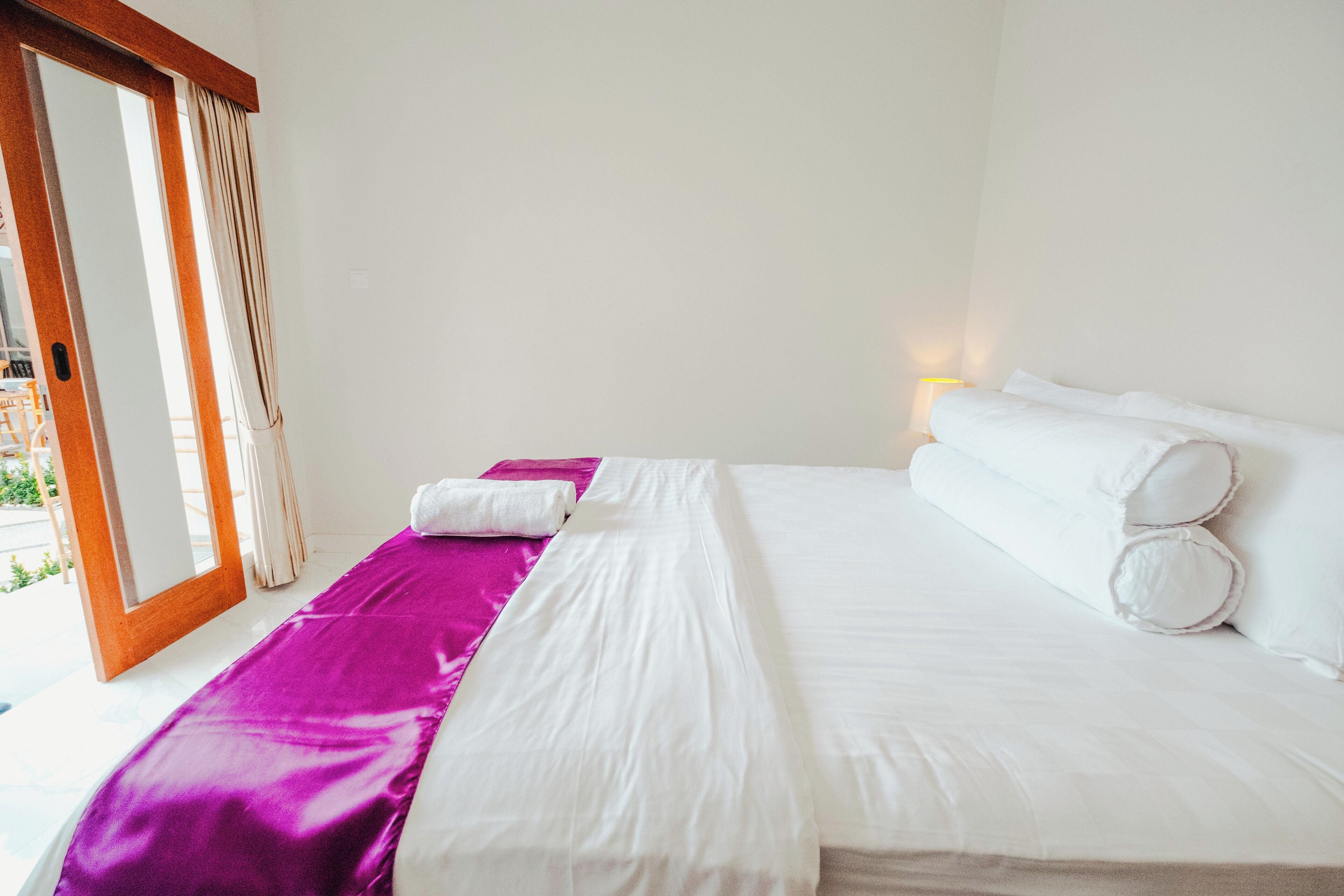 A hotel room with a bed, a purple bedspread, white pillows, and a nightstand with a lamp.