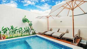A swimming pool with lounge chairs and umbrellas, surrounded by greenery and a white wall. - Turtle Shelter Guest House (Denpasar)