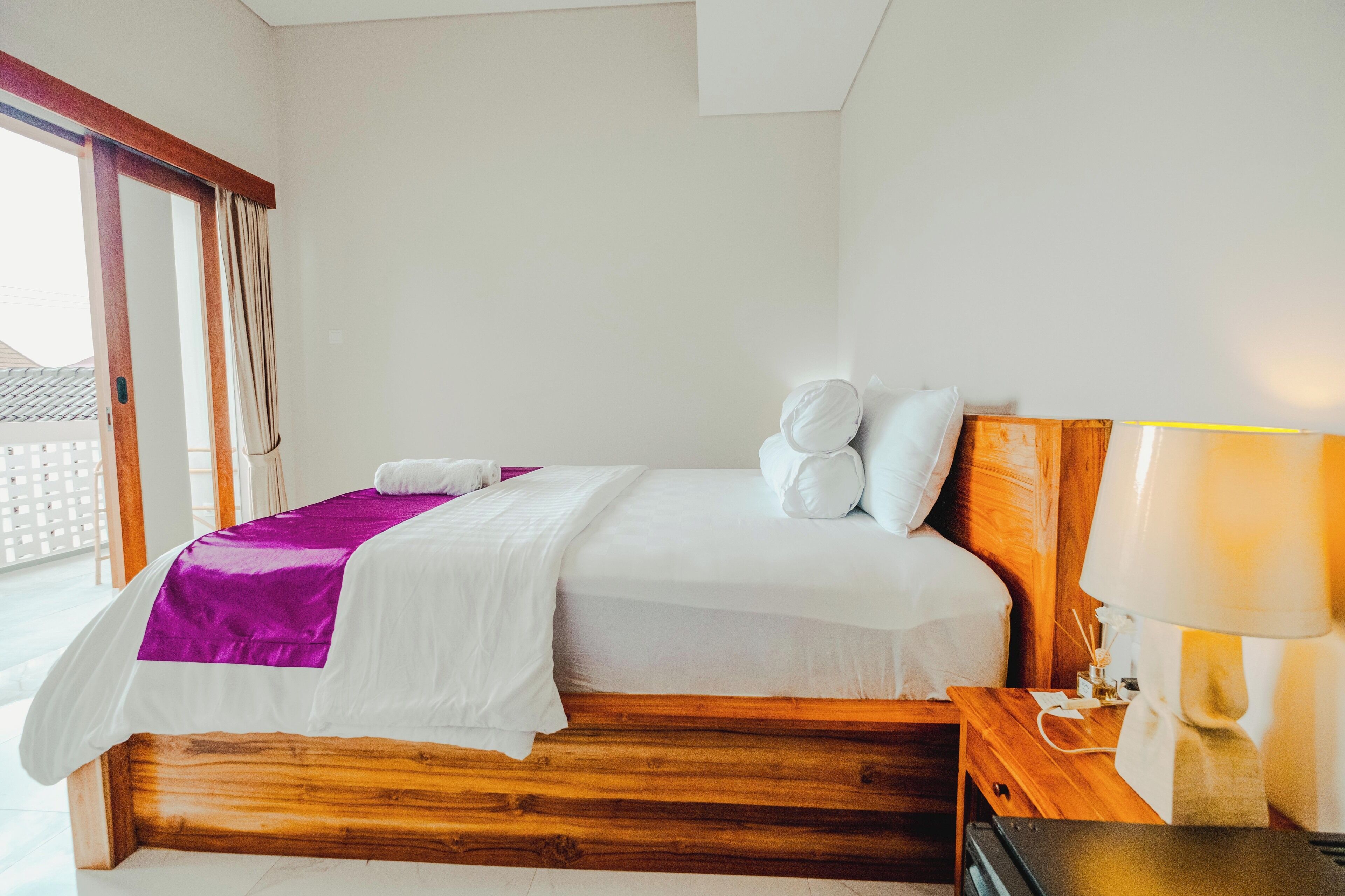 A neatly made bed with white and purple bedding, a wooden headboard, and a bedside lamp.