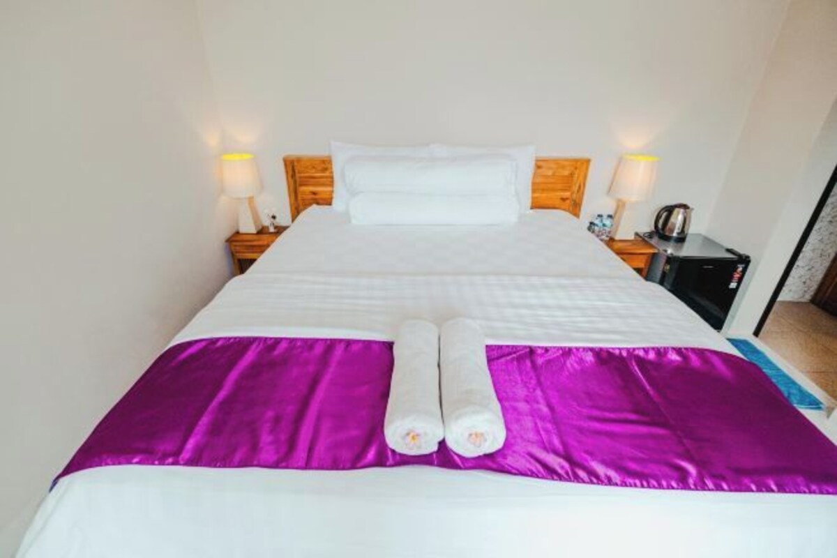 A neatly made bed with white linens and a purple bedspread, two rolled towels, and a nightstand with a lamp and a kettle.