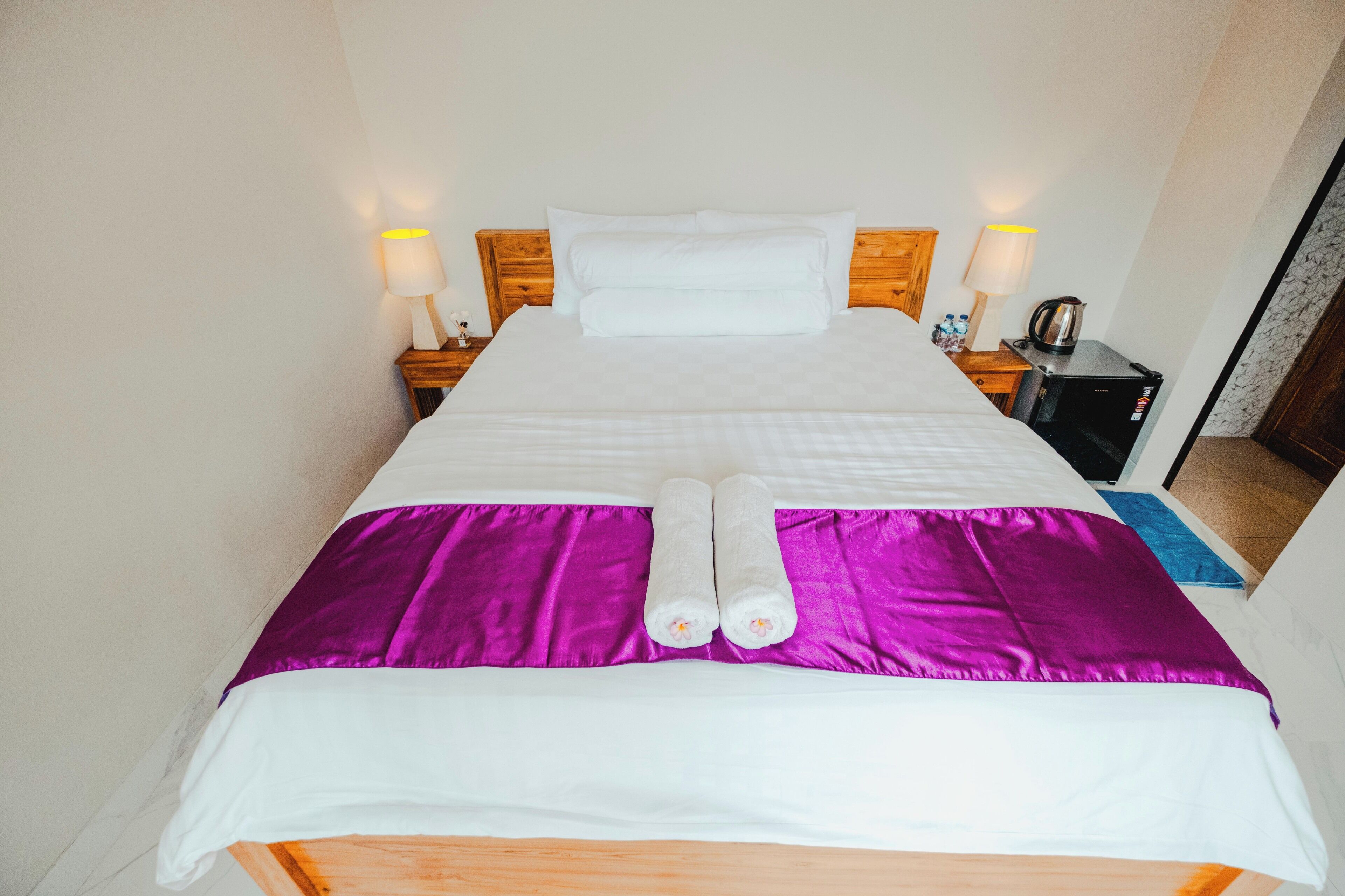 A neatly made bed with white and purple linens, two rolled towels, and bedside lamps.