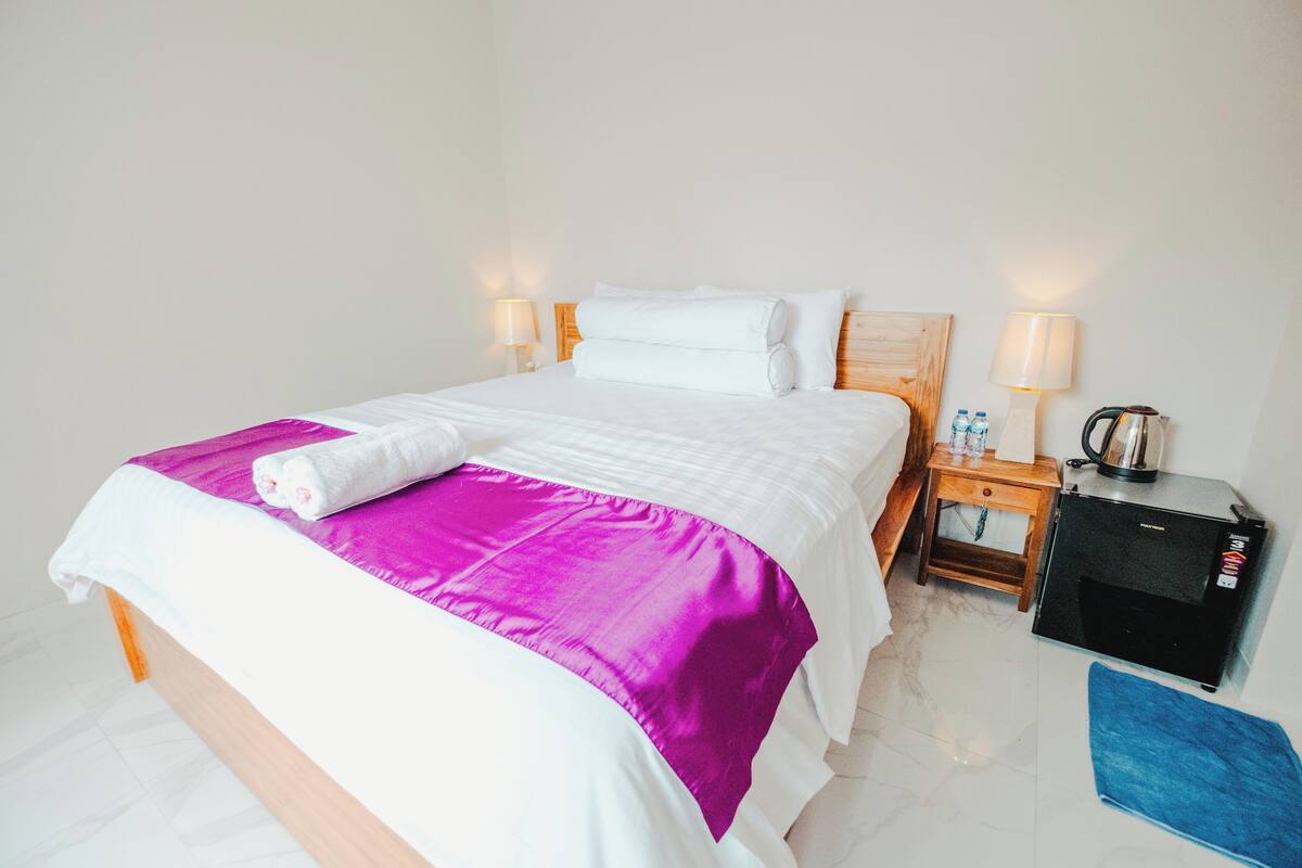 A neatly made bed with a white and purple bedspread, two bedside lamps, a small wooden nightstand with a kettle, and a black refrigerator.