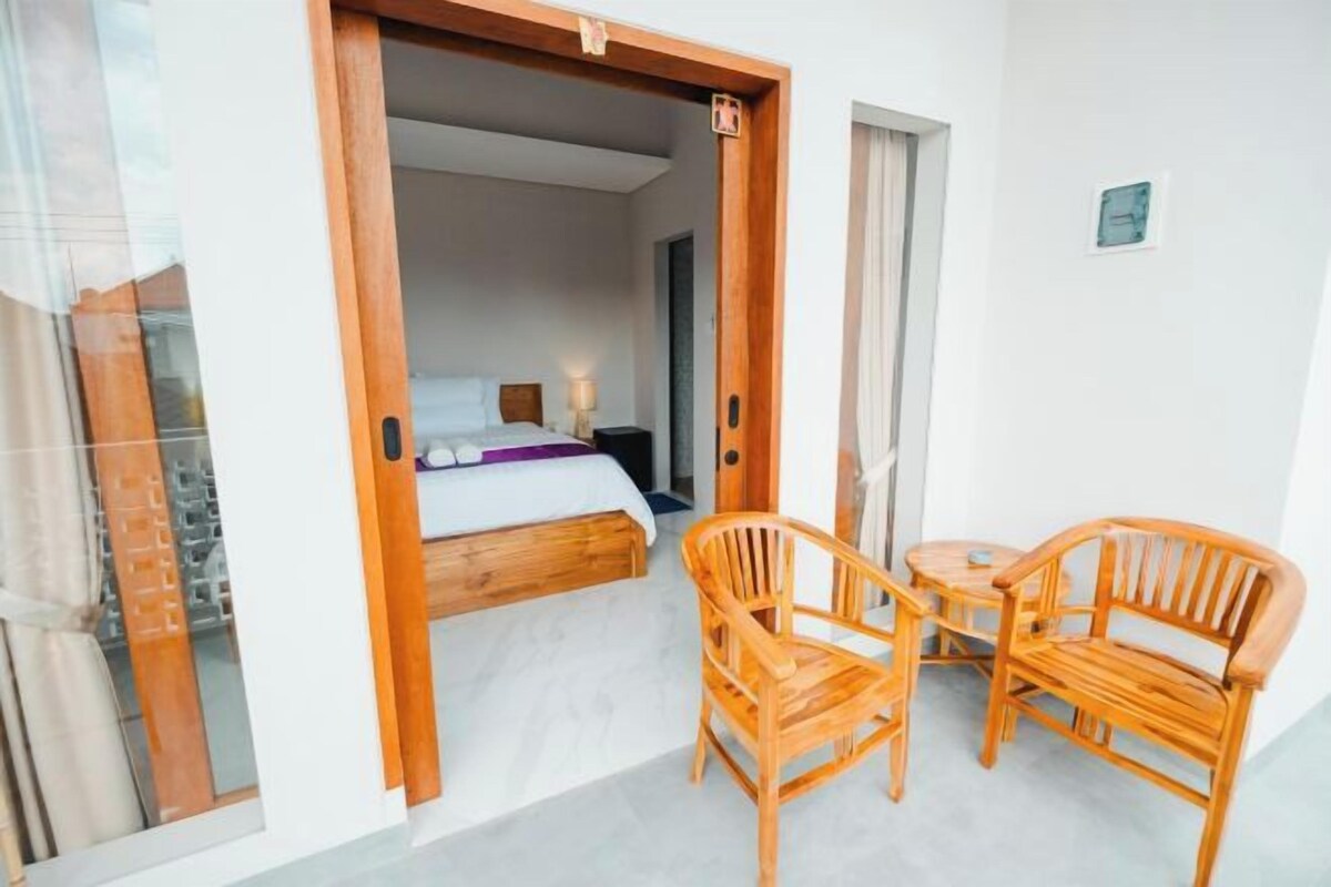 A room with a bed, a wooden chair, a small table, and a sliding glass door leading to a balcony.