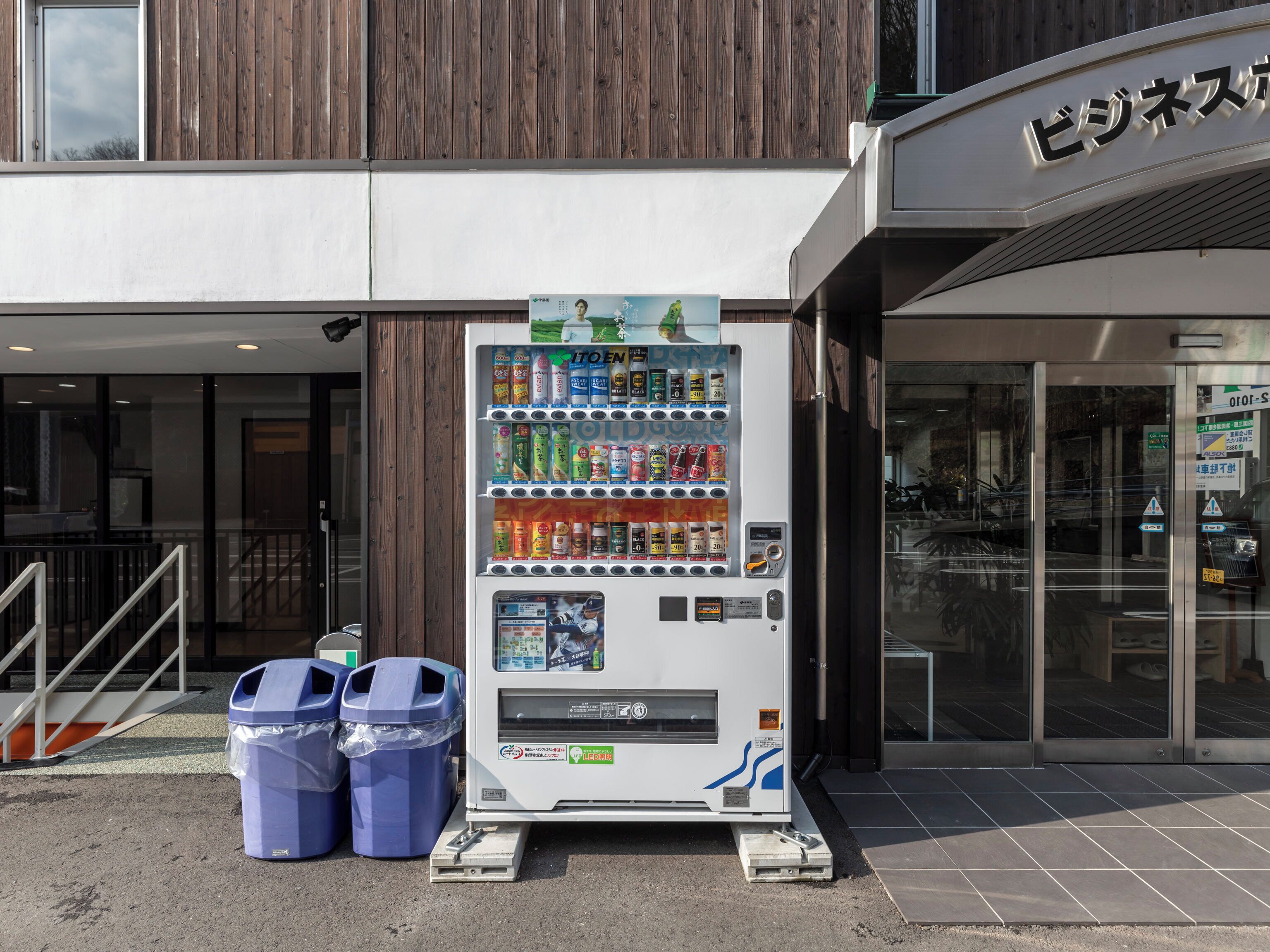 A vending machine with a variety of drinks and snacks, two blue trash bins, and a building entrance with a sign.