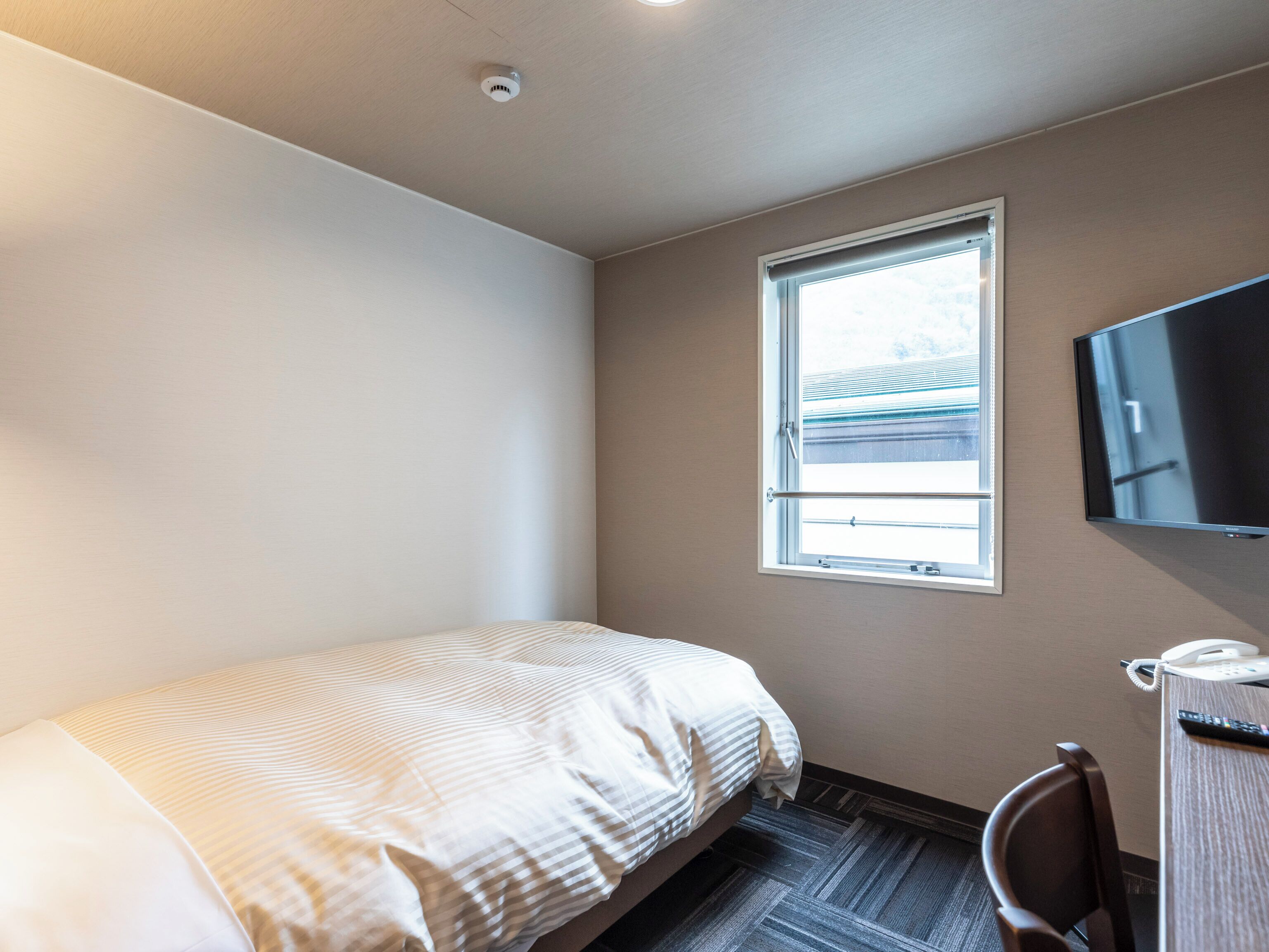 Ein Hotelzimmer mit einem Einzelbett, einem an der Wand befestigten Fernseher, einem Fenster mit Ausblick und einem Schreibtisch mit Telefon und Fernbedienung.