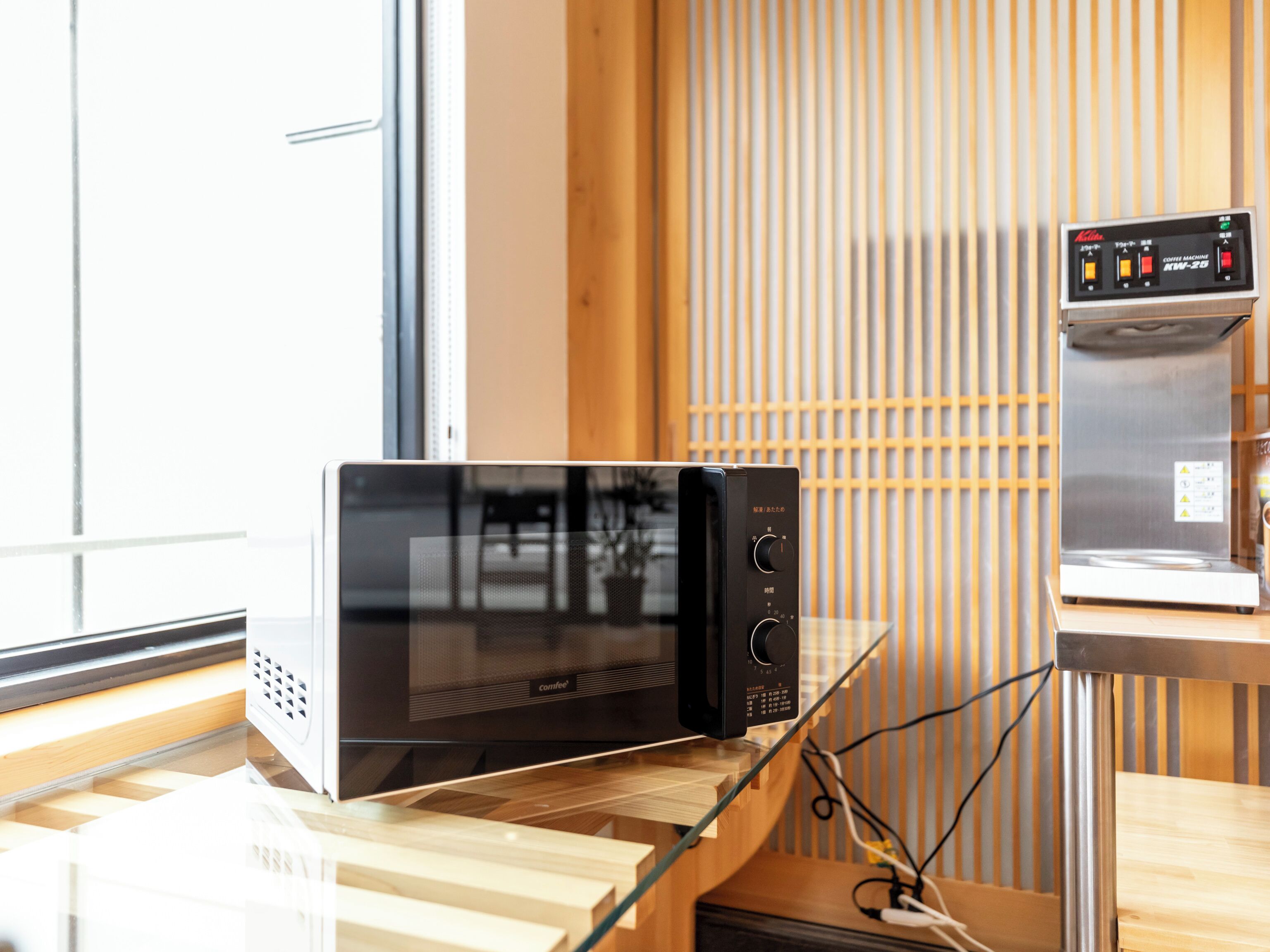 A modern kitchen with a black microwave oven on a wooden countertop, a coffee machine on a separate counter, and a window with blinds in the background.