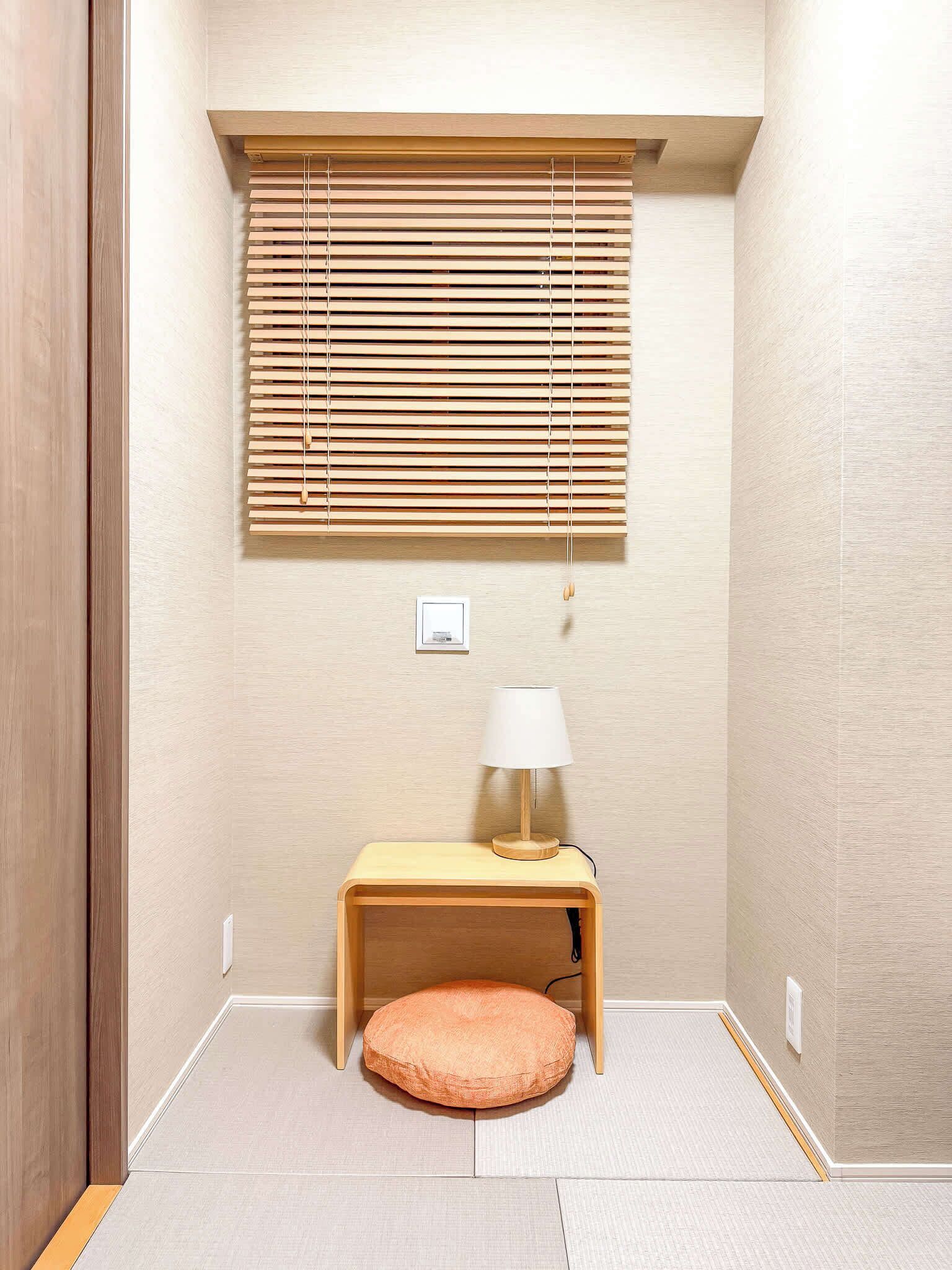A small, cozy room with a wooden floor, a round cushion, a small table with a lamp, and a window with wooden blinds.