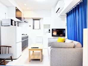 A compact living space with a kitchenette, a sofa with a yellow pillow, a television, and a blue curtain. - Triphome Sugamo 2024 New 3 mins 2 stations 25 (Toshima City)