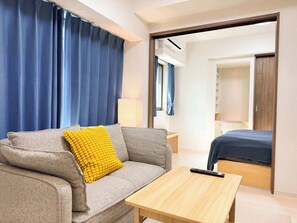 A modern hotel room with a grey sofa, a yellow pillow, a wooden table, and a bed with a blue cover. - Triphome Sugamo 2024 New 3 mins 2 stations 25 (Toshima City)