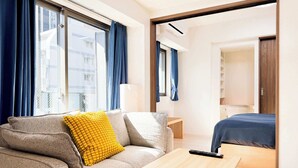 Ein modernes Hotelzimmer mit einer Couch, einem kleinen Holztisch, einem Bett und Blick auf die Stadt durch große Fenster.
