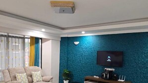 A modern living room with a beige sofa set, a glass coffee table, a blue textured accent wall, a flat-screen TV, and a wooden entertainment unit. - Shawmut Suites Nakuru (Nakuru)