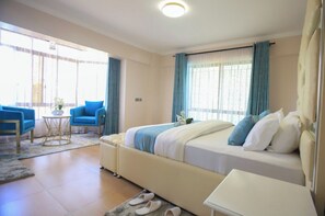 A hotel room with a large bed, a small table, a sofa, and a window with curtains. - Shawmut Suites Nakuru (Nakuru)
