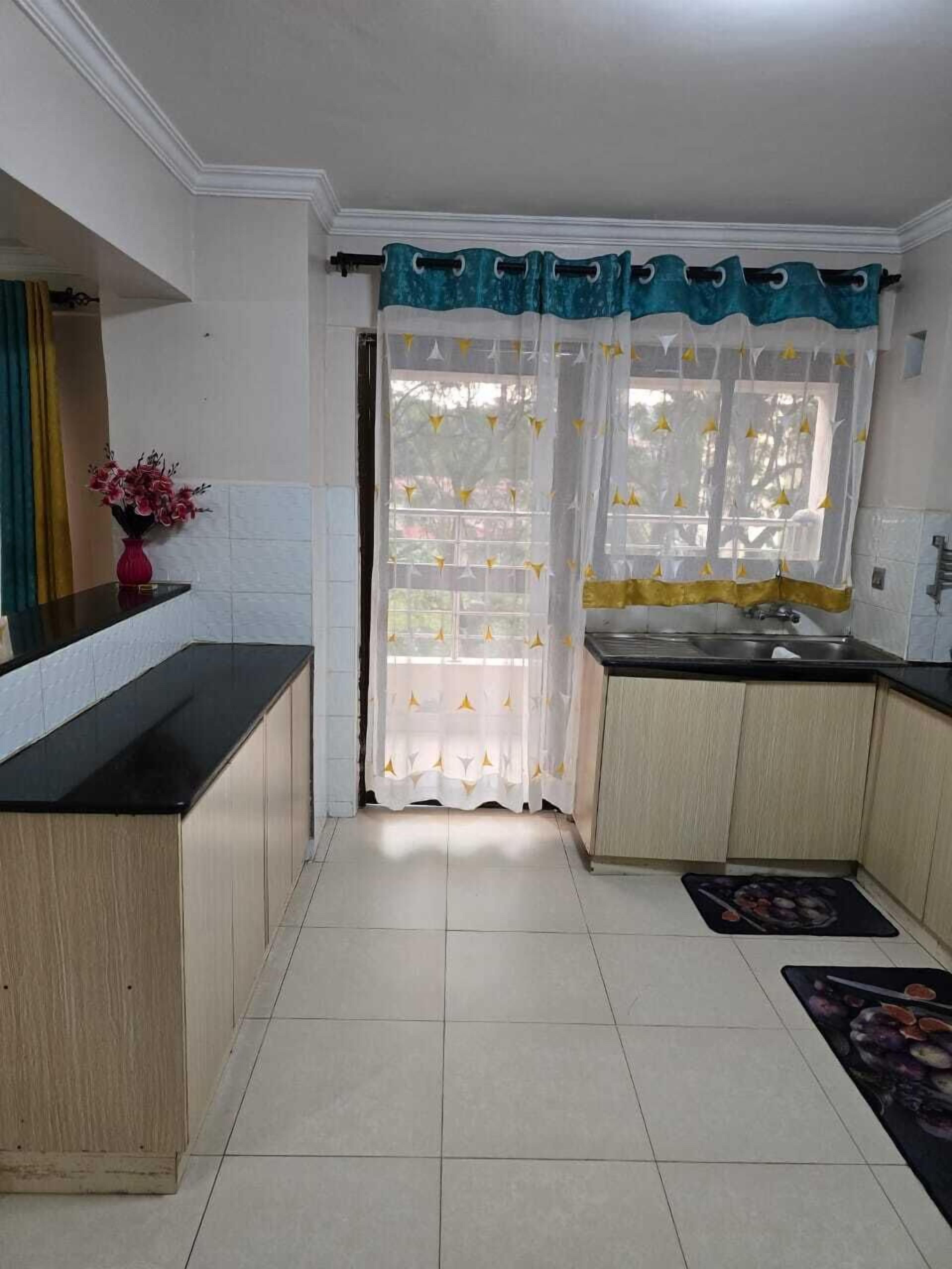 A kitchen with a black countertop, wooden cabinets, a white tiled floor, a sink, and a window with a curtain.