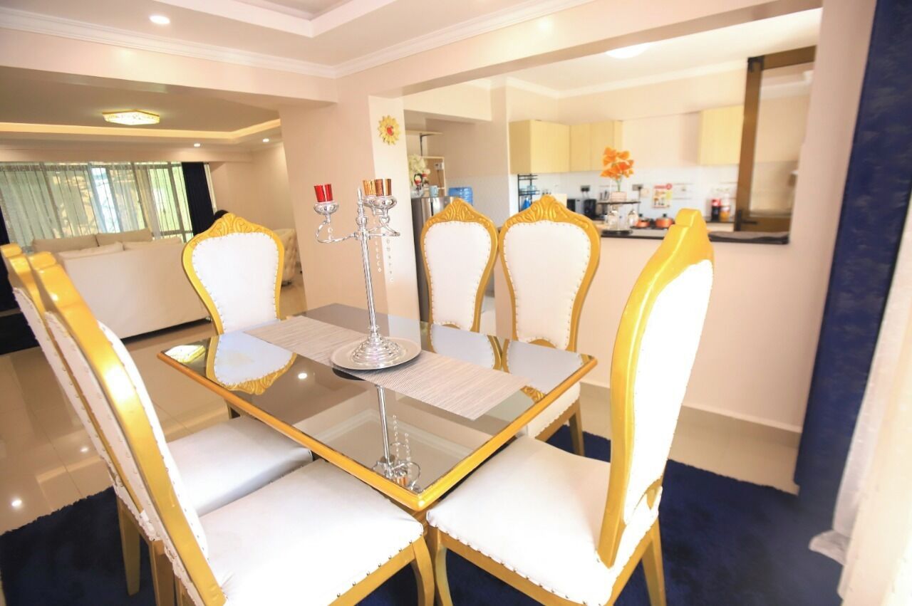 A dining area with a glass table and gold chairs, a chandelier, and a view of the kitchen in the background.
