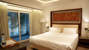 A hotel room with a large bed, a wooden nightstand, a desk with a tray, and a sliding glass door leading to a balcony. - Lords Hrim Akshara Resort (Mudhol)