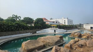 A swimming pool with large rocks, a white building in the background, and lush greenery. - Lords Hrim Akshara Resort (Mudhol)