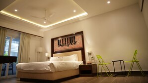 A modern hotel room with a large bed, a ceiling fan, recessed lighting, a painting above the bed, and a view of the outdoors through a sliding door. - Lords Hrim Akshara Resort (Mudhol)