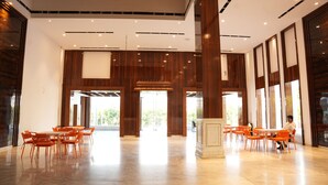 A spacious lobby with high ceilings, wooden paneling, and a central column. Tables and chairs are arranged for seating. - Lords Hrim Akshara Resort (Mudhol)