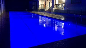 Una piscina con un borde iluminado en azul, una escalera que lleva a un nivel superior y un edificio con ventanas amplias.