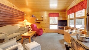 Een gezellig interieur van een blokhut met een bank, fauteuil, televisie, houten kasten en een raam met uitzicht op bomen.